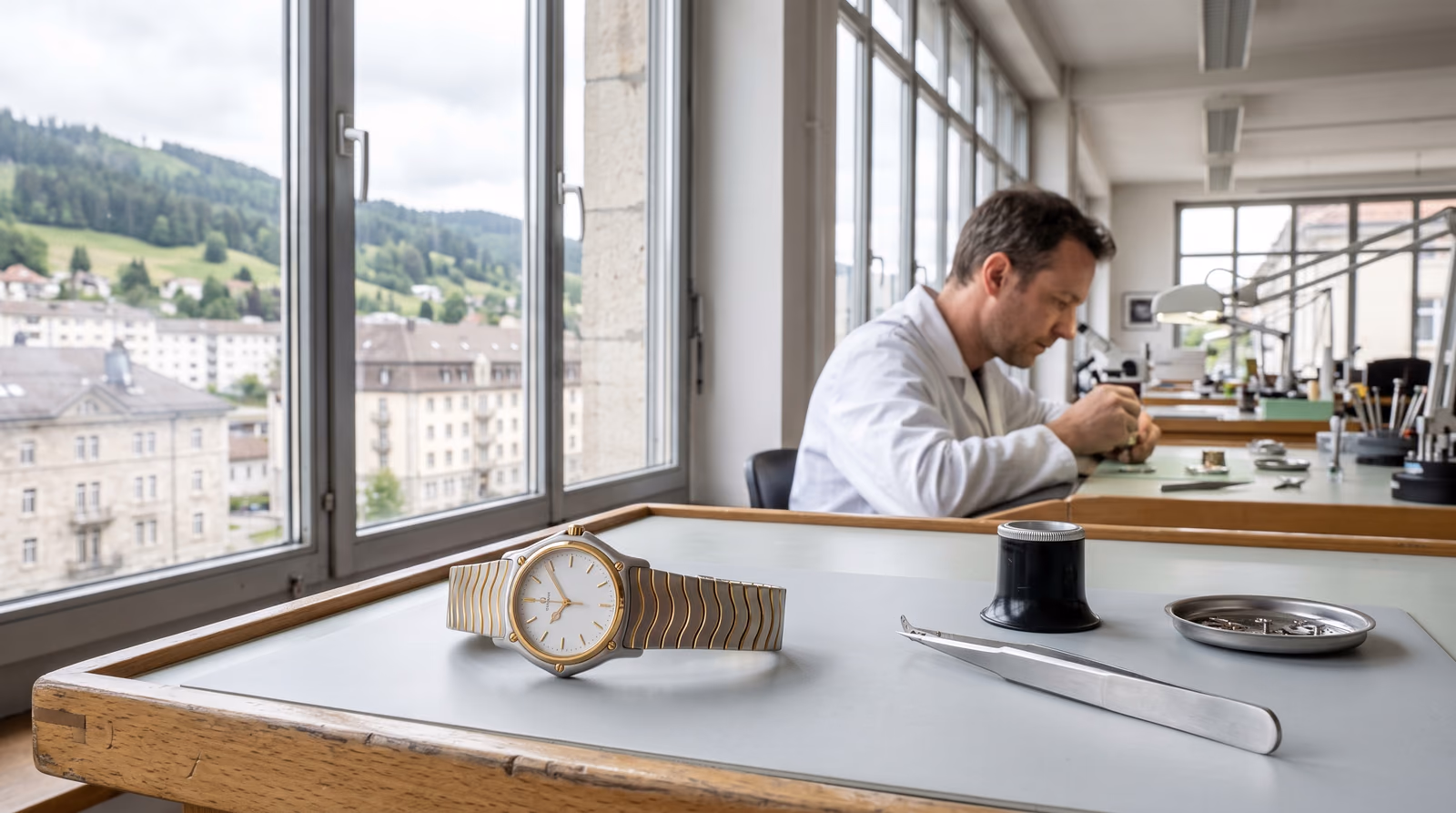 A two-tone steel and gold sports watch with a screwed bezel and wave-link bracelet lies on a watchmaker's bench beside a loupe and tweezers, while a watchmaker in a white coat works at the next bench in a bright workshop overlooking the town of La Chaux-de-Fonds