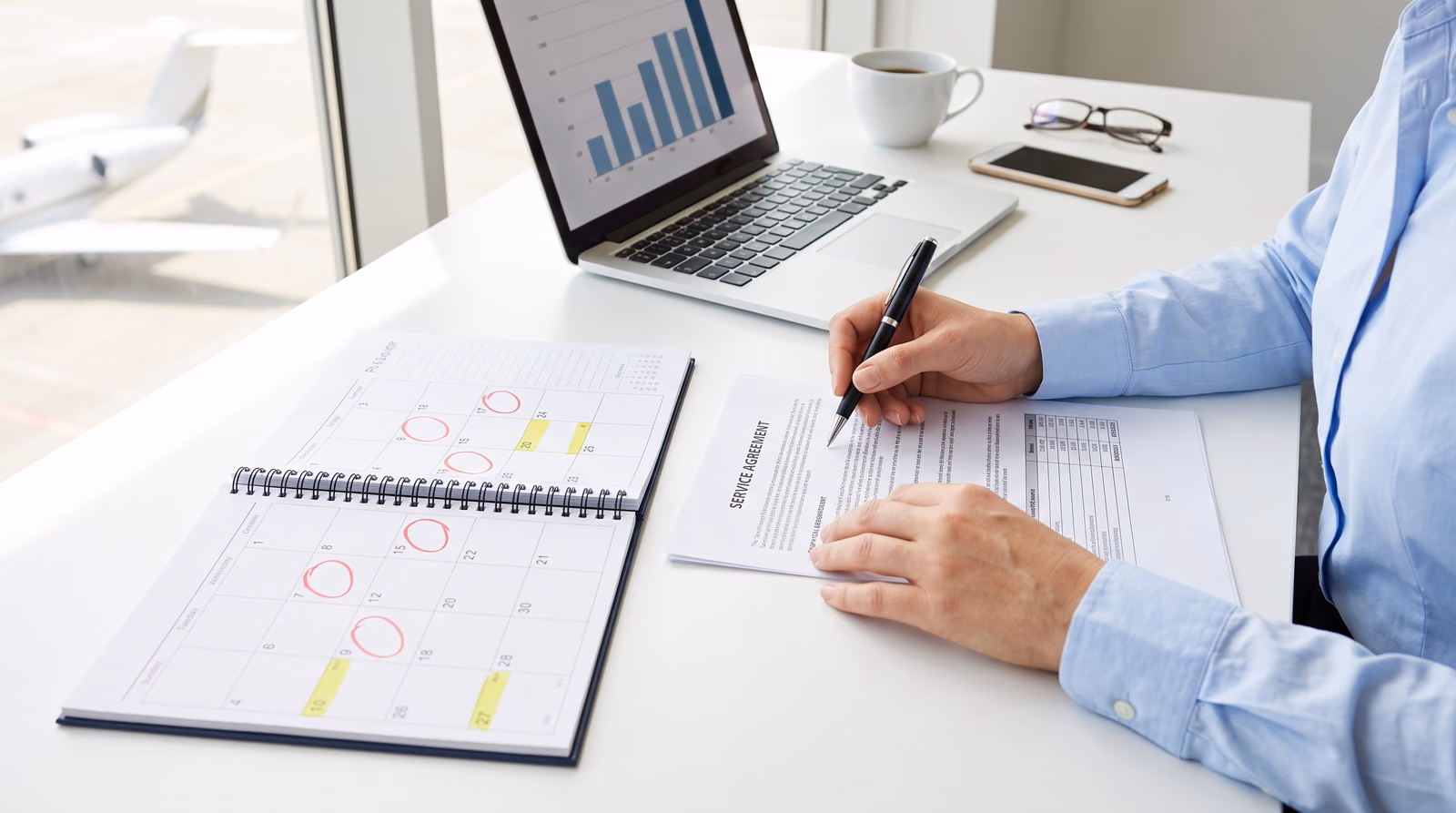 A buyer's hands with a pen over a printed service agreement on a bright white desk, beside an open desk planner with several dates circled in red and highlighted in yellow, a laptop showing a bar chart, and a business jet parked outside the window