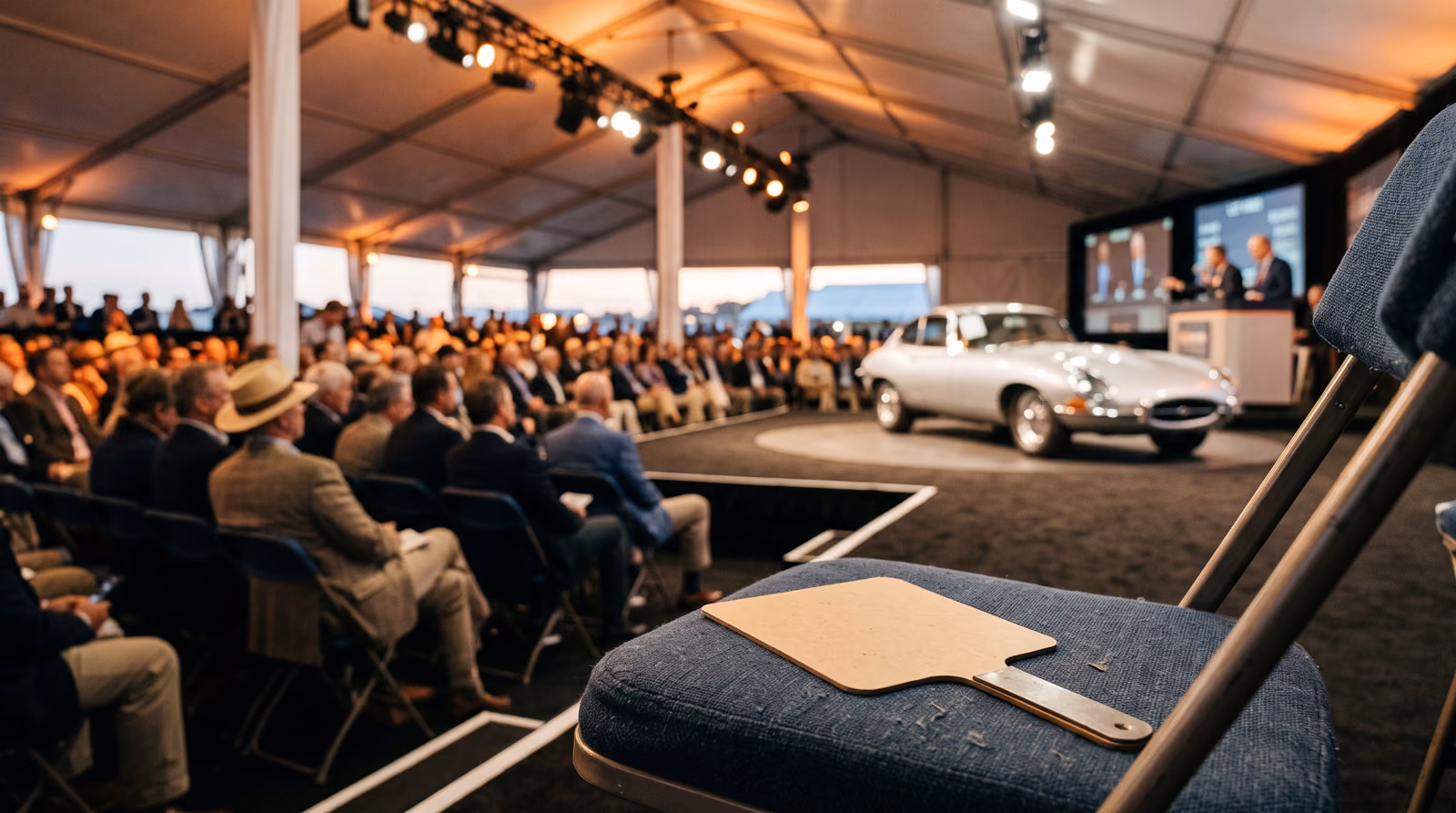 A blank cardstock bidder paddle lying face down on an empty folding chair at the front of a crowded collector car auction marquee, a silver 1960s coupe on the rostrum behind under rigged stage lighting