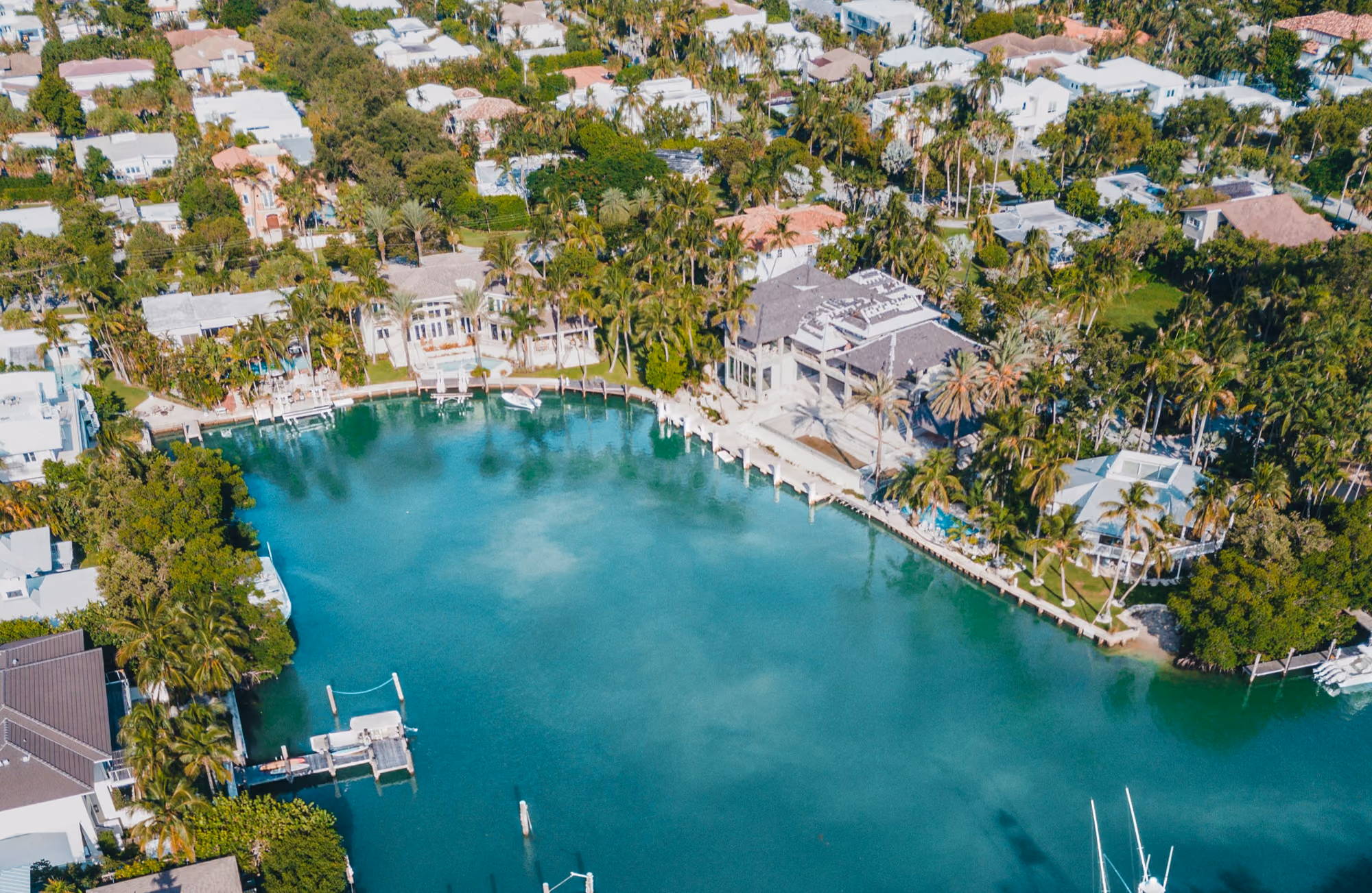 Aerial view of a serene waterfront neighborhood with lush trees, white houses, and boats docked along turquoise water under a sunny sky.