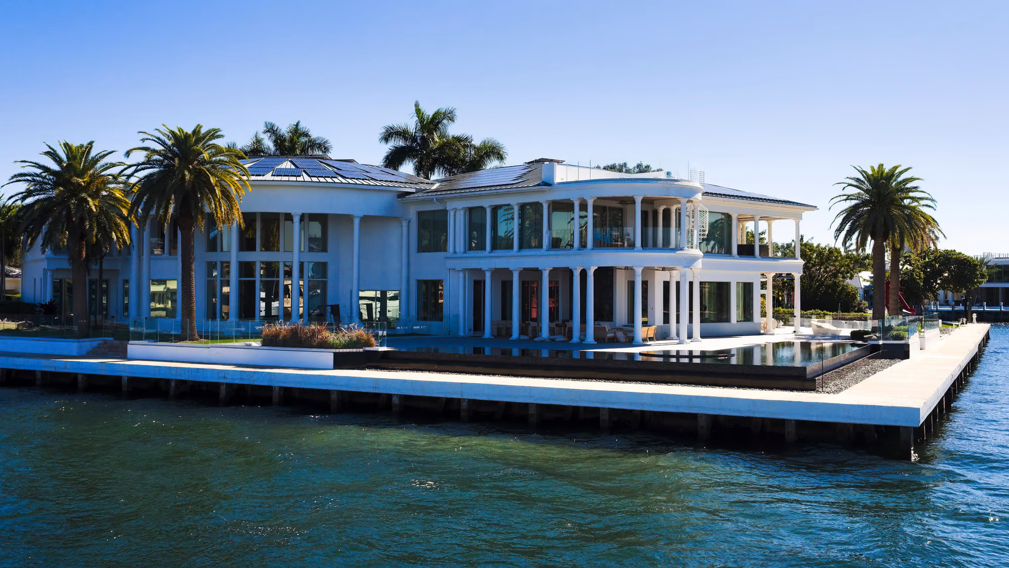 Luxurious white waterfront mansion with palm trees, surrounded by blue waters. Modern architecture, clear skies, serene atmosphere.