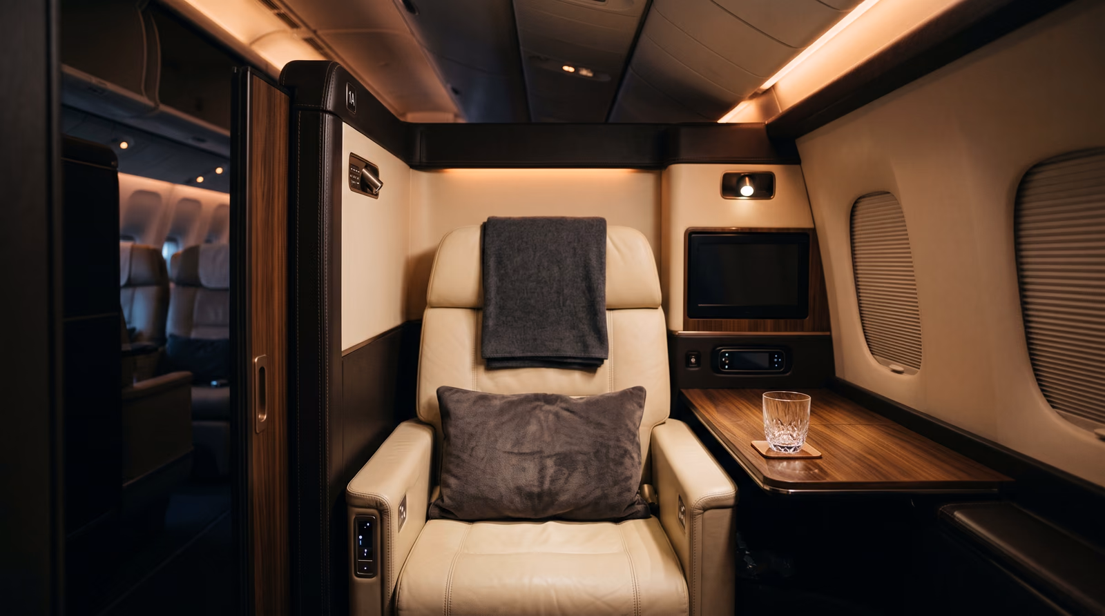 An empty premium airline suite with a cream leather seat, folded throw and side table under warm cabin lighting.