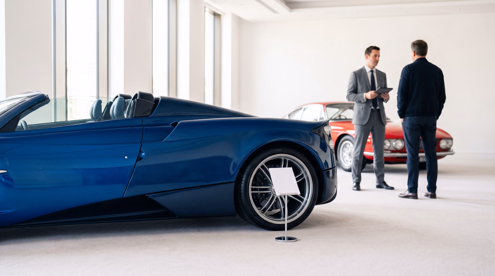 A deep blue open-top supercar with exposed carbon fibre bodywork stands beside a blank lot card in a bright, daylit auction preview room, while an auction specialist holding a tablet talks with a visitor in front of a red 1960s Italian coupe