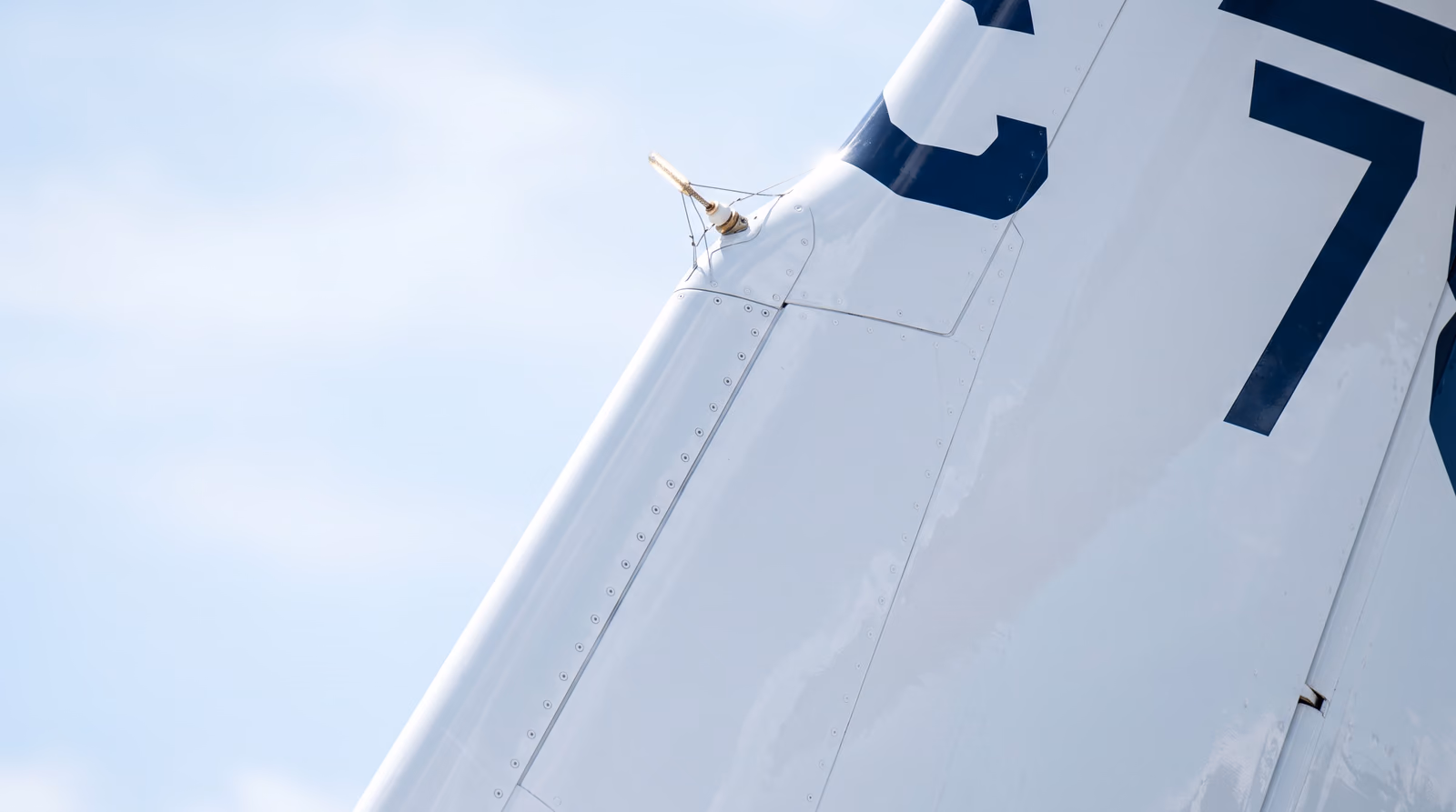 Tight daylight close-up of the white vertical stabiliser of a business jet against a pale blue sky, with fragments of dark navy painted registration characters cropped by the edge of the frame and a static wick on the fin tip.